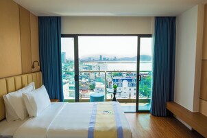A hotel room with a bed, a view of a cityscape, a small table with a plant, and a bench.