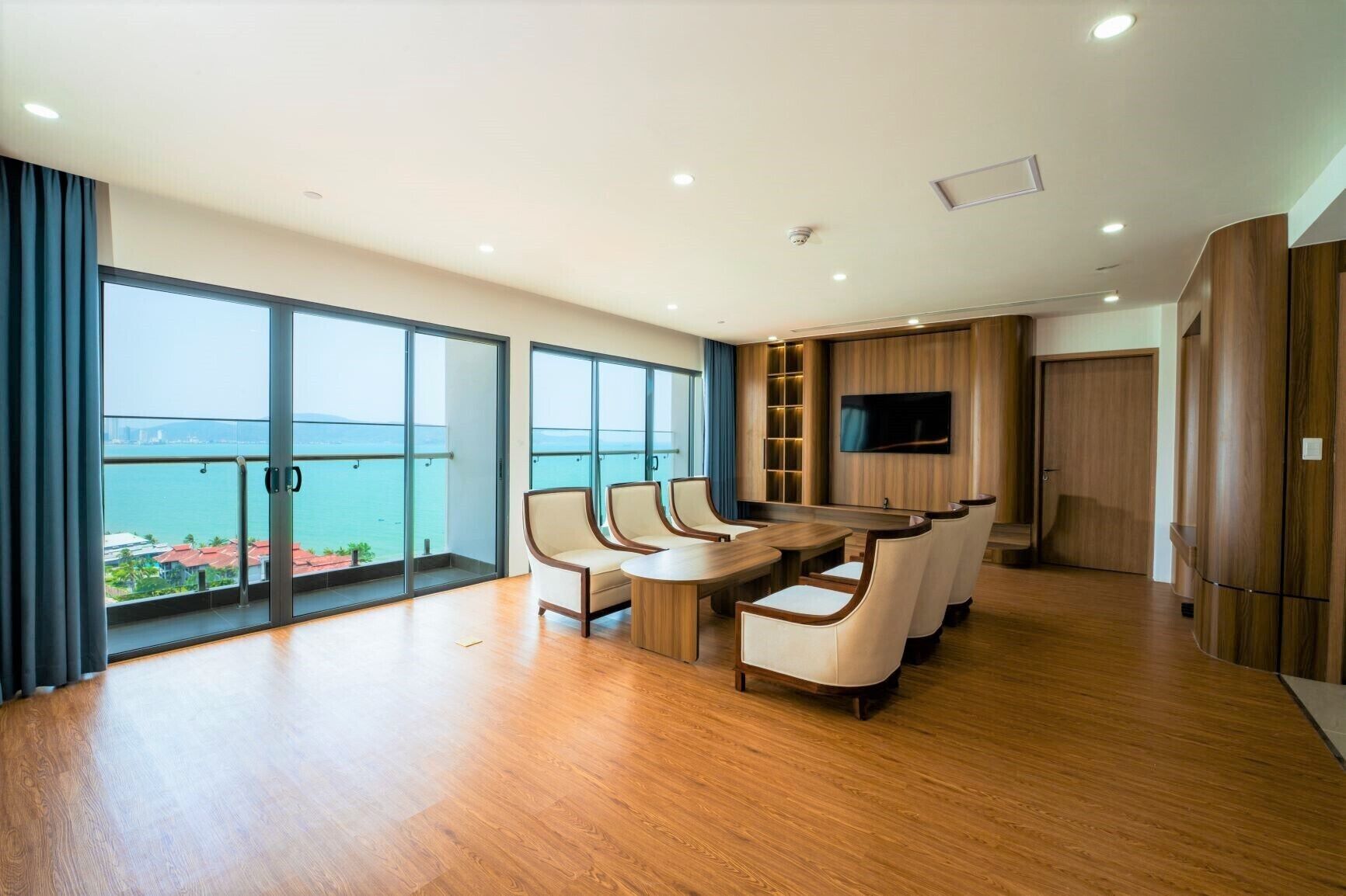 A spacious room with a large dining table and chairs, a flat-screen TV mounted on the wall, and a balcony with a view of the sea.