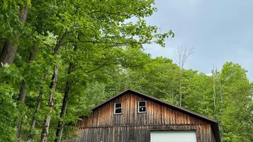 Une grange rustique en bois, avec une porte de garage blanche, entourée d’arbres verdoyants et bordée d’un chemin de gravier qui y mène.