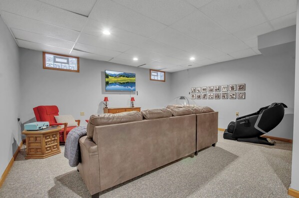 Un salon avec un canapé beige, un fauteuil rouge, un fauteuil massant, une table d’appoint en bois et un téléviseur fixé au mur.