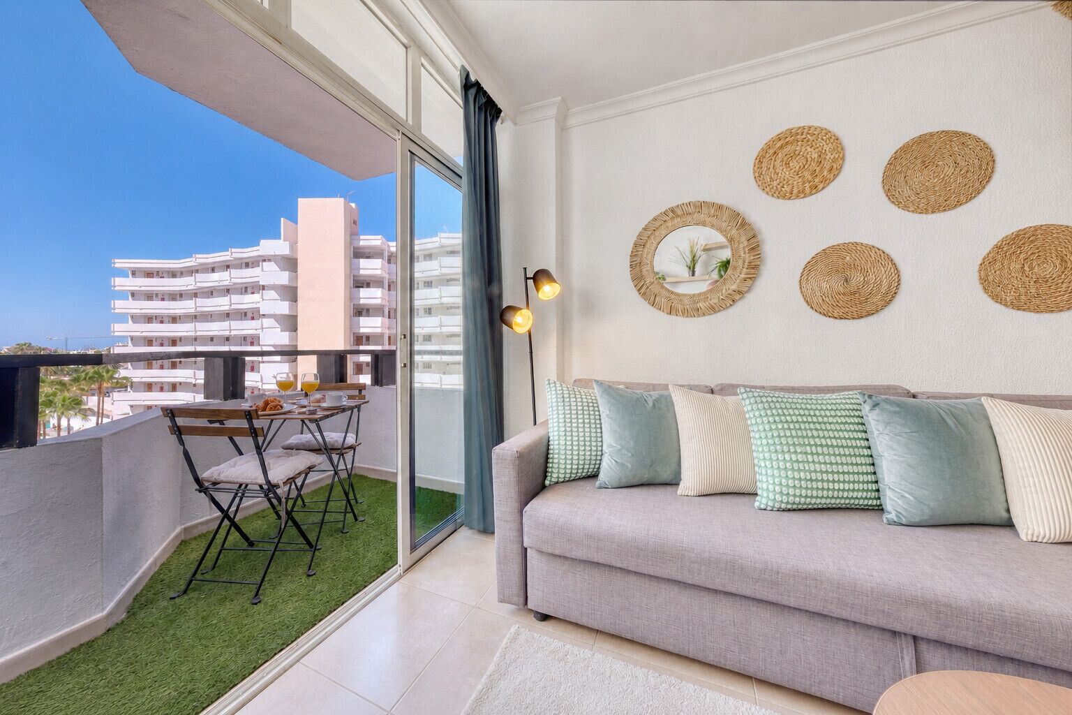 A balcony with a table and chairs, a grey sofa with patterned cushions, and a wall with woven circular decorations.
