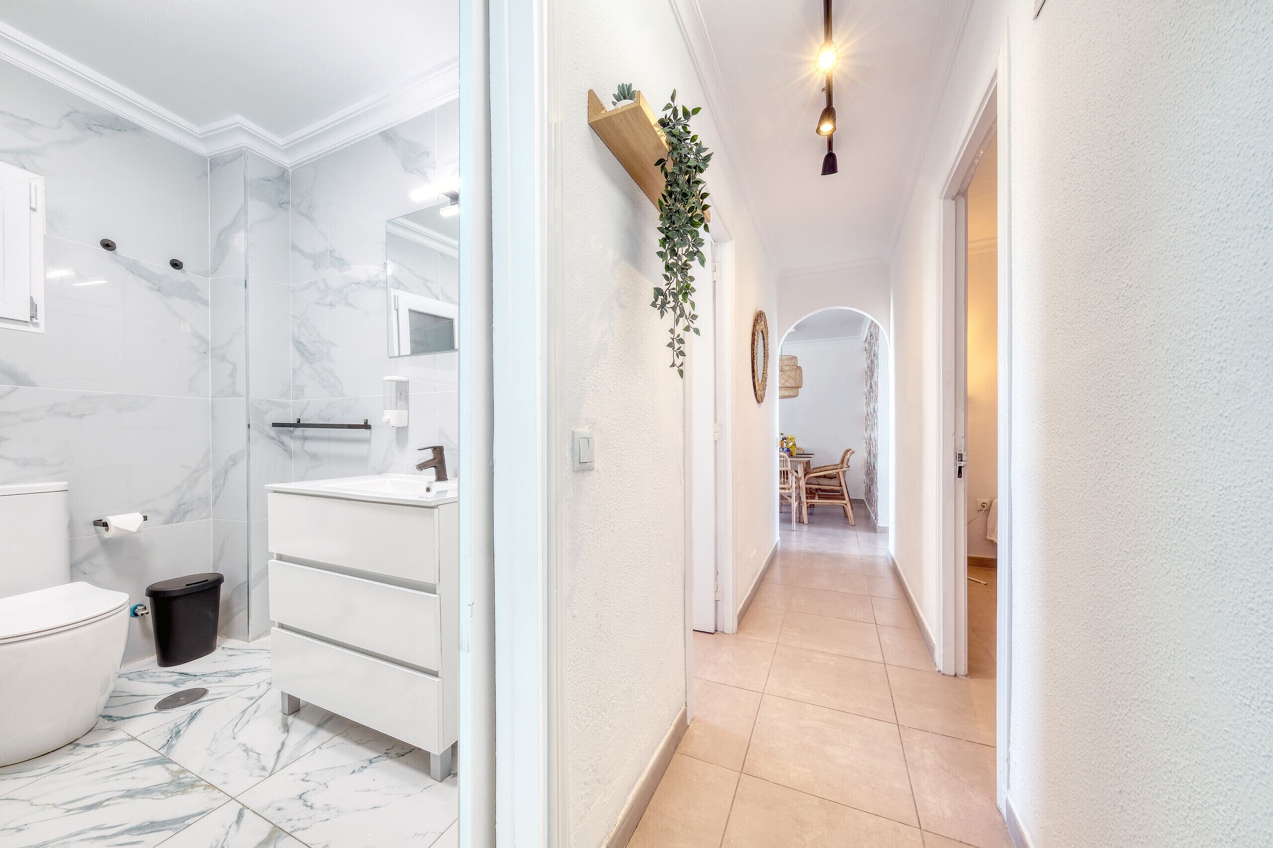 Une salle de bain avec des toilettes, un lavabo et un miroir, ainsi qu’un couloir agrémenté d’une plante et d’une porte donnant sur une autre pièce.