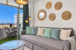 Ein modernes Wohnzimmer mit einem grauen Sofa, grünen und beigen Kissen, einem runden Spiegel und Wandbehängen aus gewebtem Material.