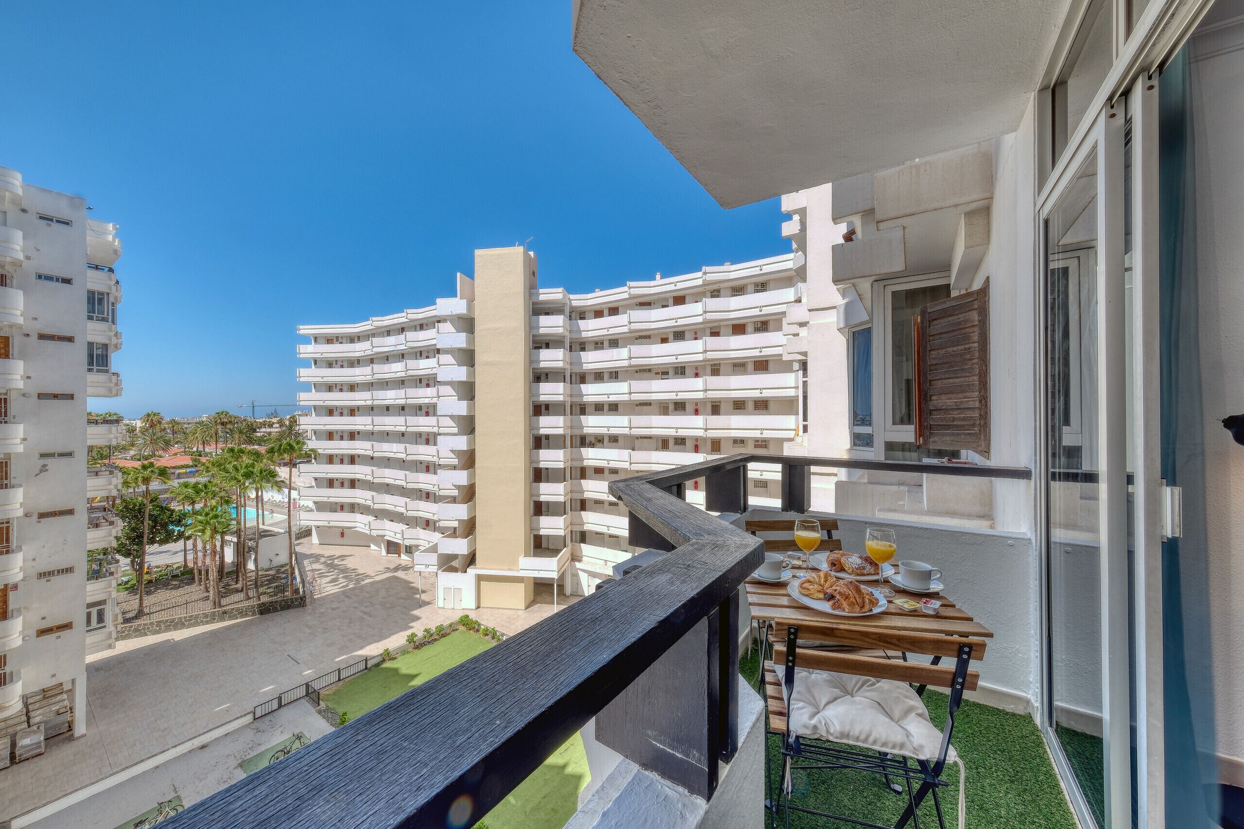 Un balcón con una mesa lista para el desayuno, vista a un edificio residencial y un cielo azul despejado.