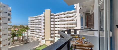 Un balcón con una mesa lista para el desayuno, vista a un edificio residencial y un cielo azul despejado.