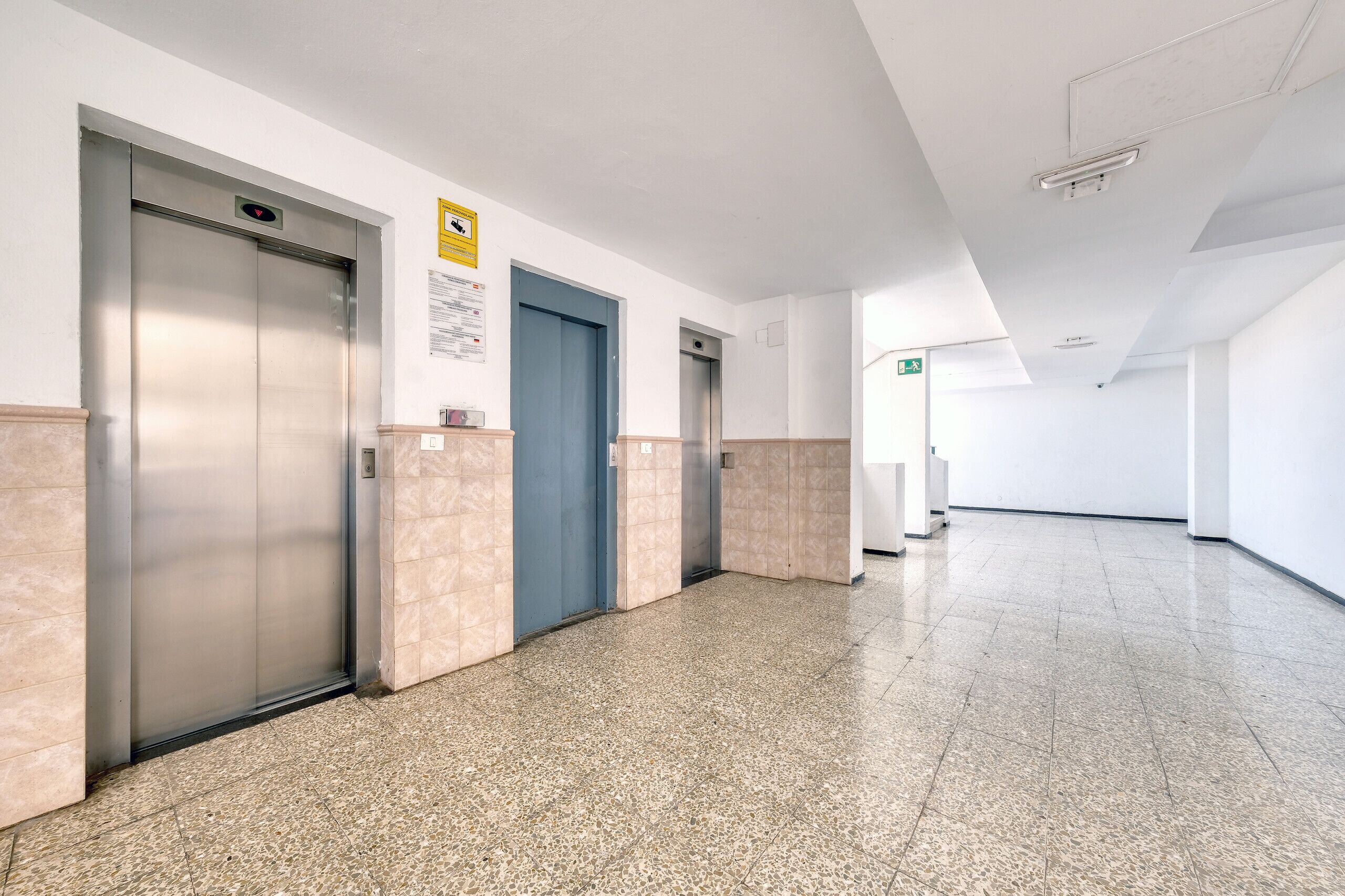 Un pasillo limpio y moderno con dos ascensores, una puerta azul y una pared blanca con un letrero amarillo.