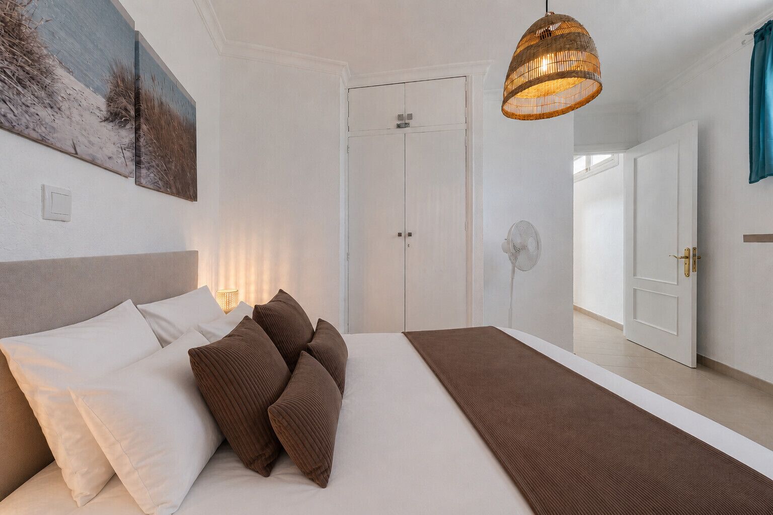 Un dormitorio con cama, almohadas, una mesita de noche, un ventilador de pared, una puerta y una lámpara colgante.