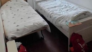 Una habitación pequeña con cama individual, un armario de madera, una ventana con cortinas y una bolsa roja en el suelo.