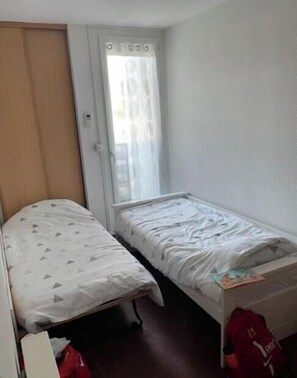 A small, single-bed room with a wooden wardrobe, a window with curtains, and a red bag on the floor. - Apartment F3 Terrace on the port of Carnon 5 minutes from the beach (Mauguio)