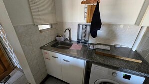 A compact kitchen with a sink, a dish rack, a washing machine, and a window with blinds. - Summer Sands Air b n b (Durban)