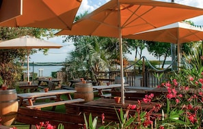 An outdoor patio with wooden tables and benches, orange umbrellas, and a view of a body of water. - Cozy (sta)caravan in Aureilhan (Aureilhan)