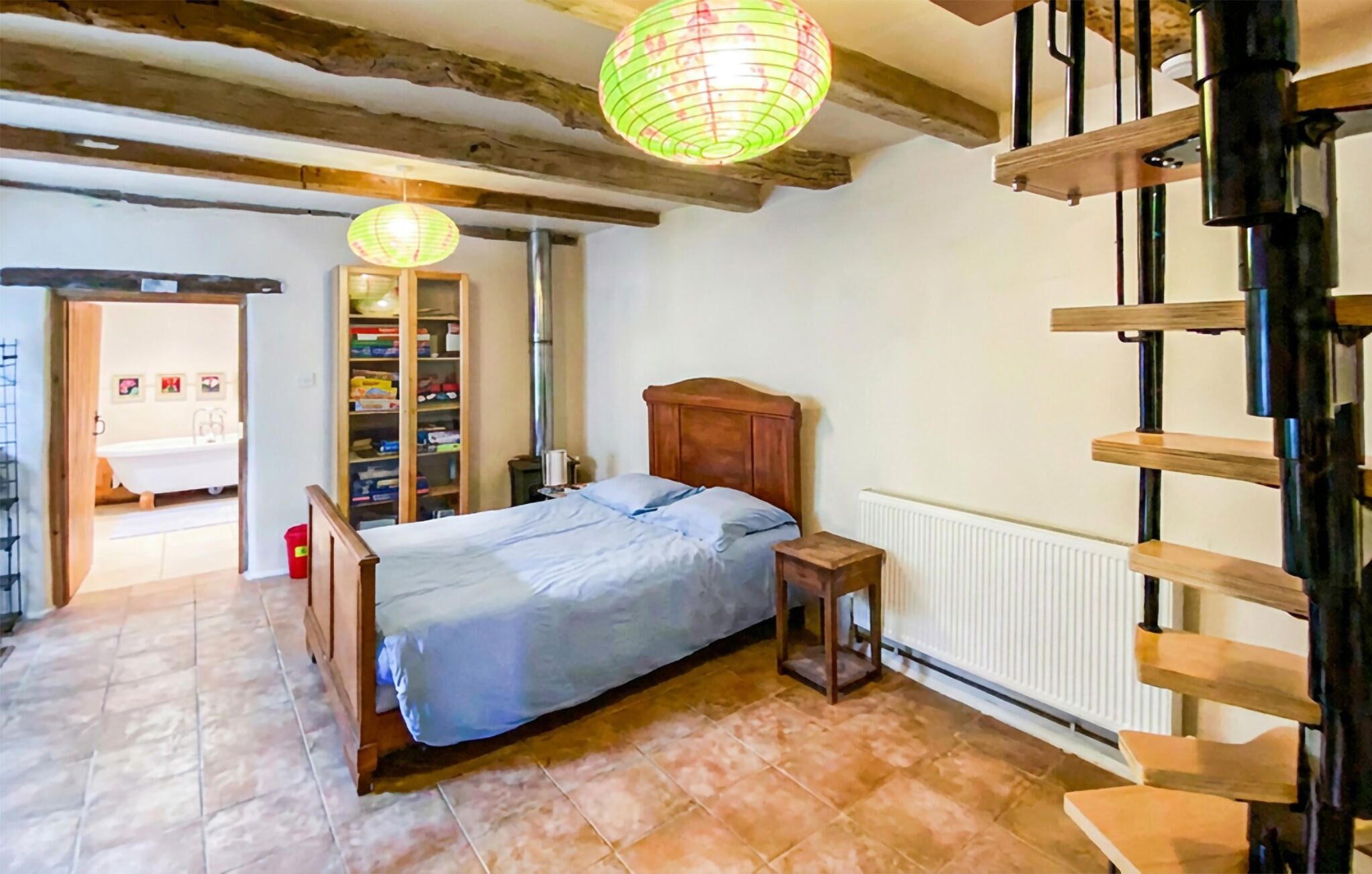 Un dormitorio con una cama de madera, una mesita de noche, un radiador, una escalera de madera y un baño visible a través de una puerta abierta.
