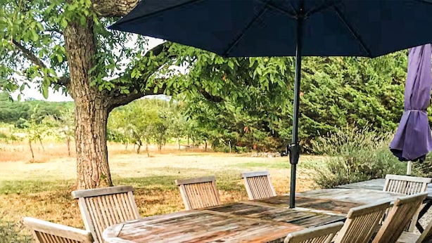Een houten tafel met stoelen en een grote parasol in een openluchtomgeving.