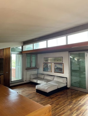 Una sala de estar moderna con suelo de madera, un sofá seccional, una mesa de comedor y ventanas grandes con contraventanas.