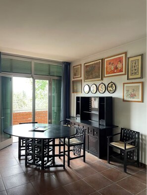 A dining area with a round table and chairs, a black cabinet with shelves, and a large window with blinds.