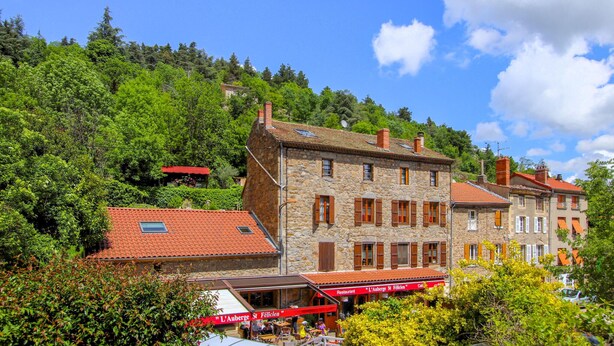 Ein Gebäude aus Stein mit rotem Ziegeldach, ein Café mit Außenbereich und üppiges Grün im Hintergrund.