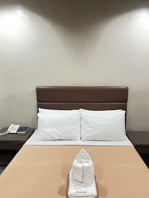 A hotel room with a bed, pillows, a folded towel, a bedside table with a phone and a book, and a nightstand.