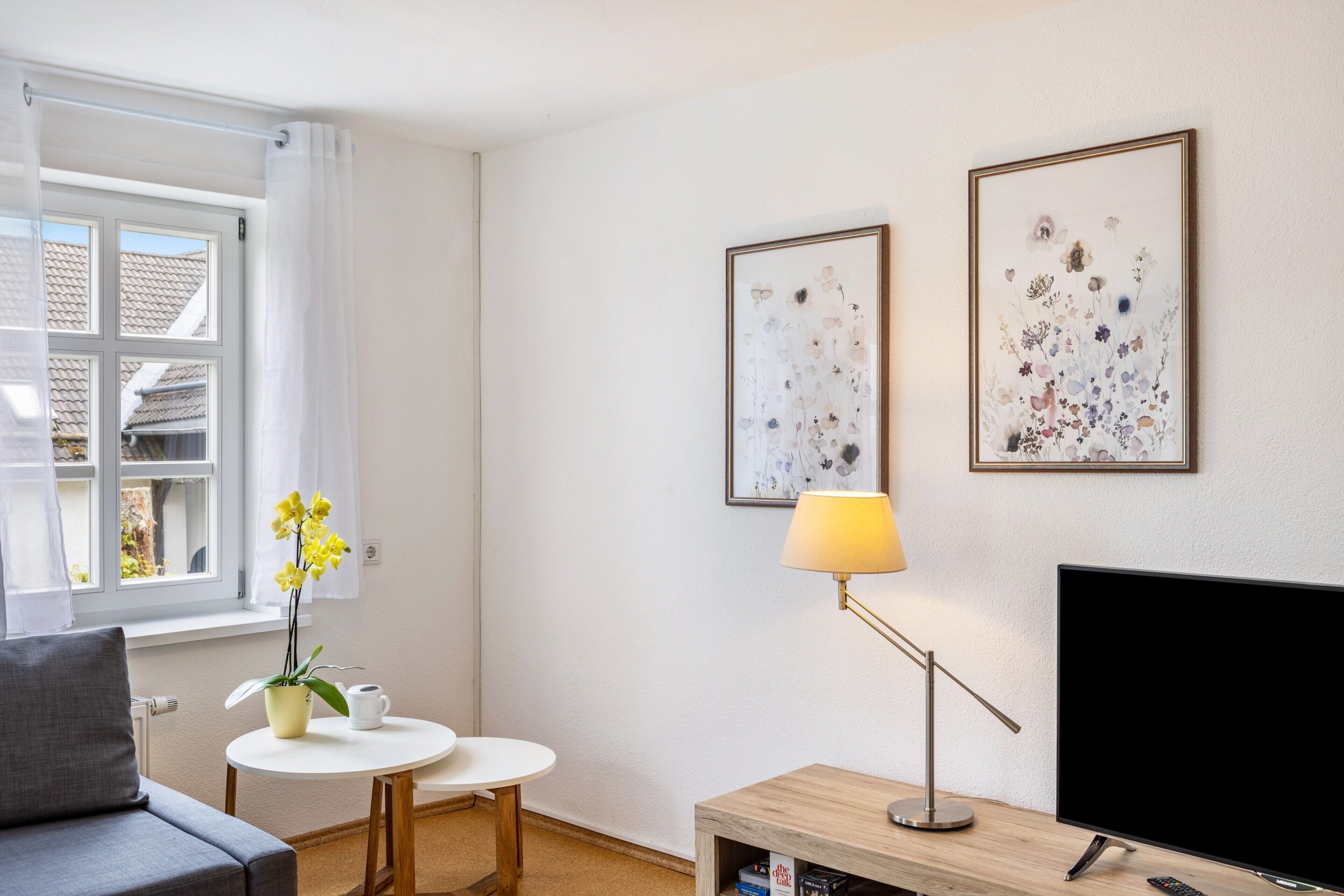 Ein modernes Wohnzimmer mit einer Couch, einem kleinen runden Tisch mit Lampe, einem Fernseher und zwei gerahmten Blumenbildern an der Wand.