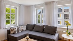 A room with a grey sofa, white curtains, and a window with a view of buildings and greenery. - Apartment 'Fewo Am Uplandsteig' with Shared Terrace, Garden and Wi-Fi (Diemelsee)