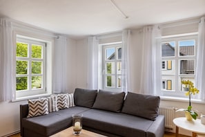 A room with a grey sofa, white curtains, and a window with a view of buildings and greenery.