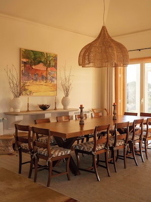 A dining room with a large wooden table, multiple chairs, a hanging light fixture, and a painting on the wall.
