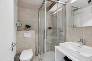 Moderni kylpyhuone, jossa on lasiseinällä varustettu suihku, valkoinen pesuallas ja wc.
