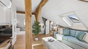A modern loft-style room with a sloped ceiling, wooden beams, a large window, a sofa, a TV, and a potted plant. - Waterfront Penthouse | AC | Rooftop | Free Parking (Zürich)