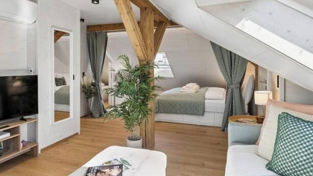 A modern loft bedroom with a sloped ceiling, a television, a sofa, a bedside table, and a window with curtains. - Waterfront Penthouse | AC | Rooftop | Free Parking (Zürich)
