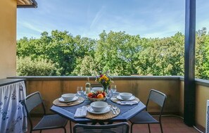 Ein Esstisch ist gedeckt mit Tellern, Schüsseln, Besteck und einer Obstschale, umgeben von Stühlen, auf einem Balkon mit Blick auf Bäume.
