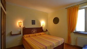 Quarto com cama, duas luminárias na parede, uma pequena pintura, um espelho redondo e janela com cortinas.