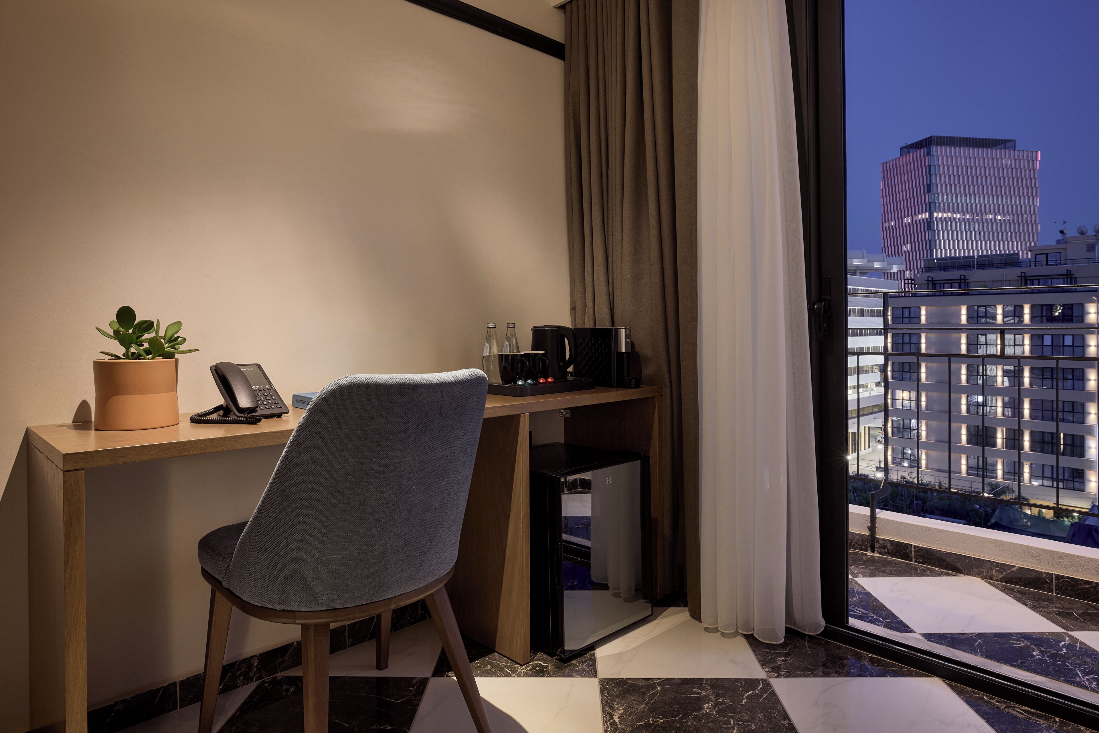 Habitación de hotel con escritorio, silla, planta en maceta, teléfono y vista panorámica de la ciudad.