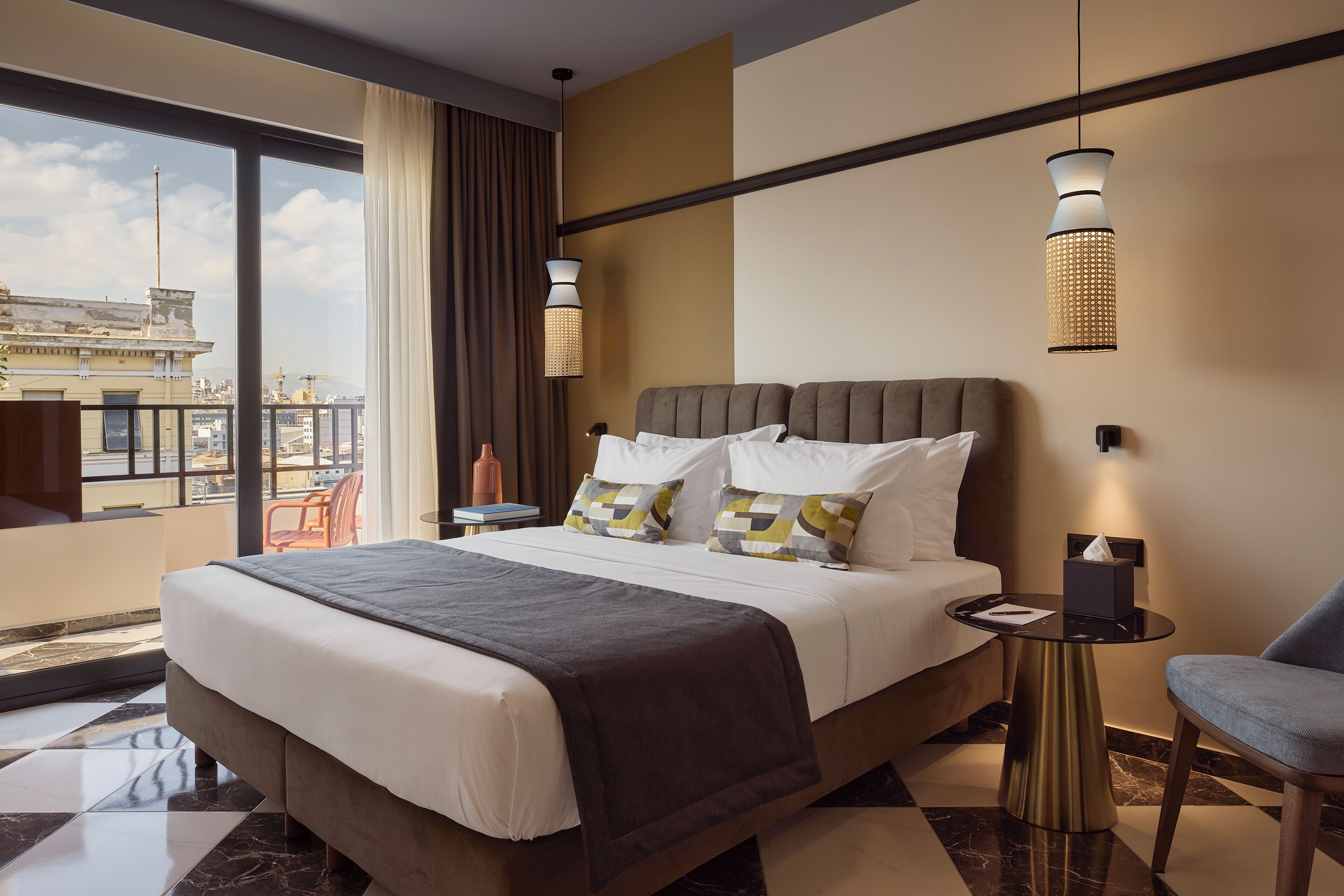 A modern hotel room with a large bed, a small table, a chair, and a view of the cityscape.