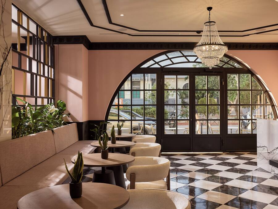 A hotel lobby with a checkered floor, seating area, large arched windows, and a chandelier.