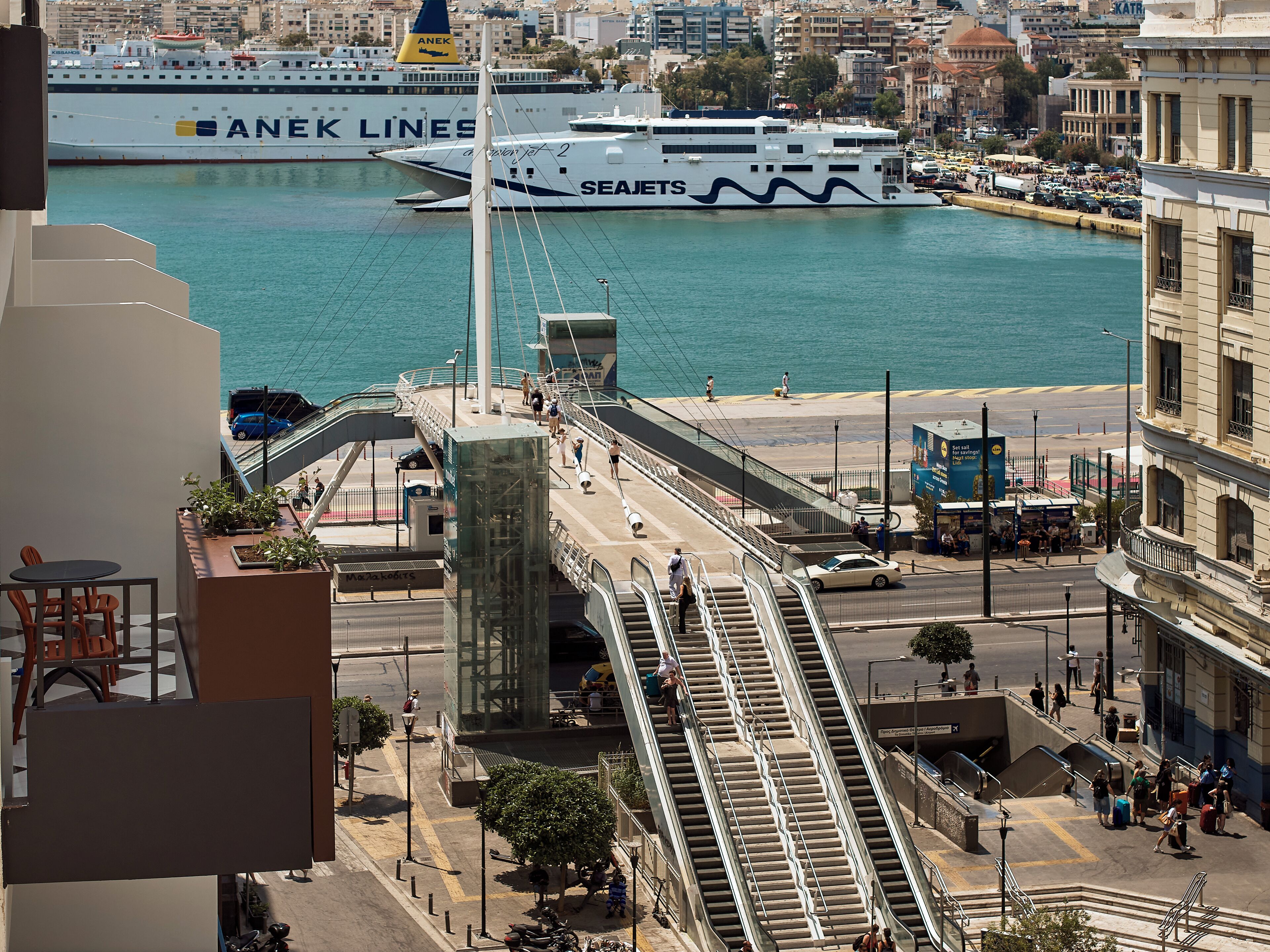 Un traghetto con la scritta 'ANEK LINES' e 'SEAJETS', un ponte pedonale e una città con edifici e un lungomare.
