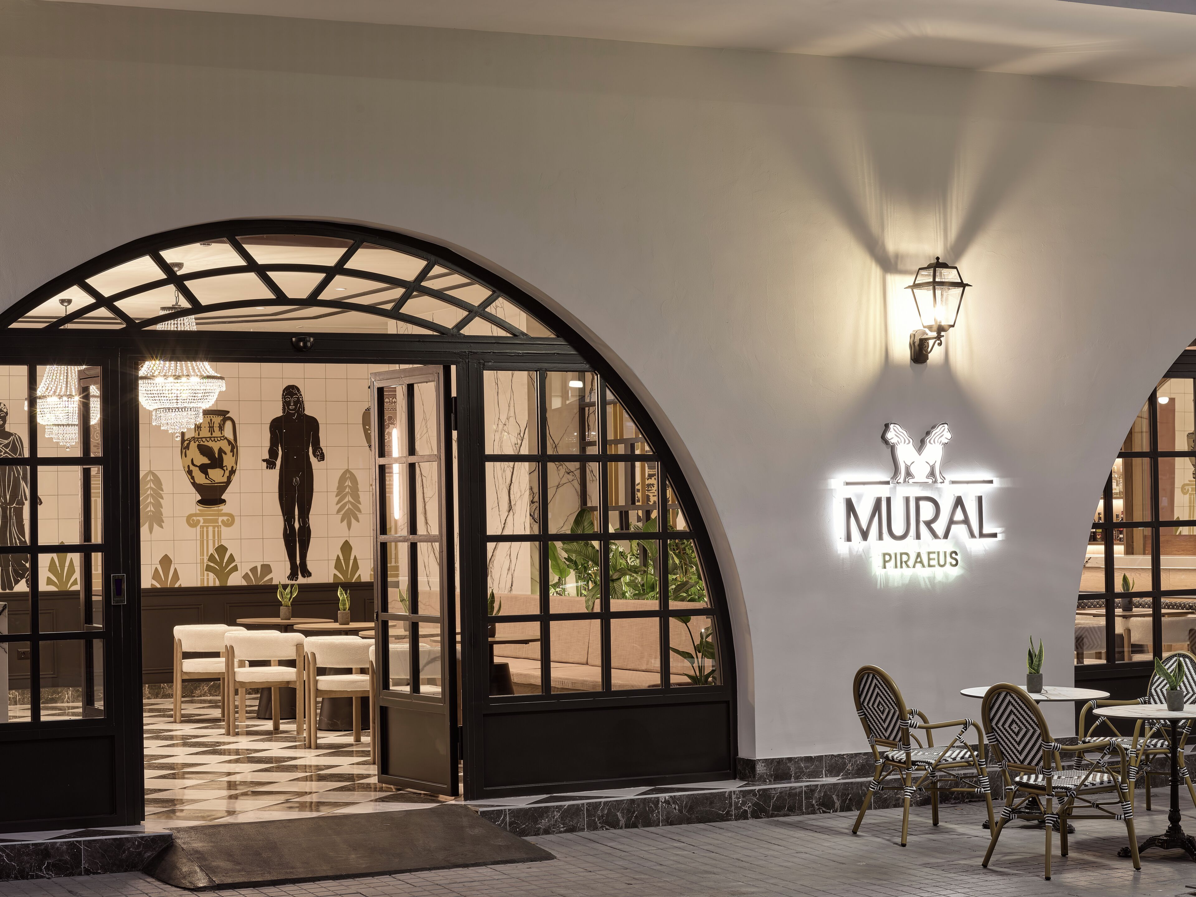 Entrada de restaurante con diseño moderno, destacando un gran arco de acceso, un área de estar con mesas y sillas, y un letrero fijado a la pared.