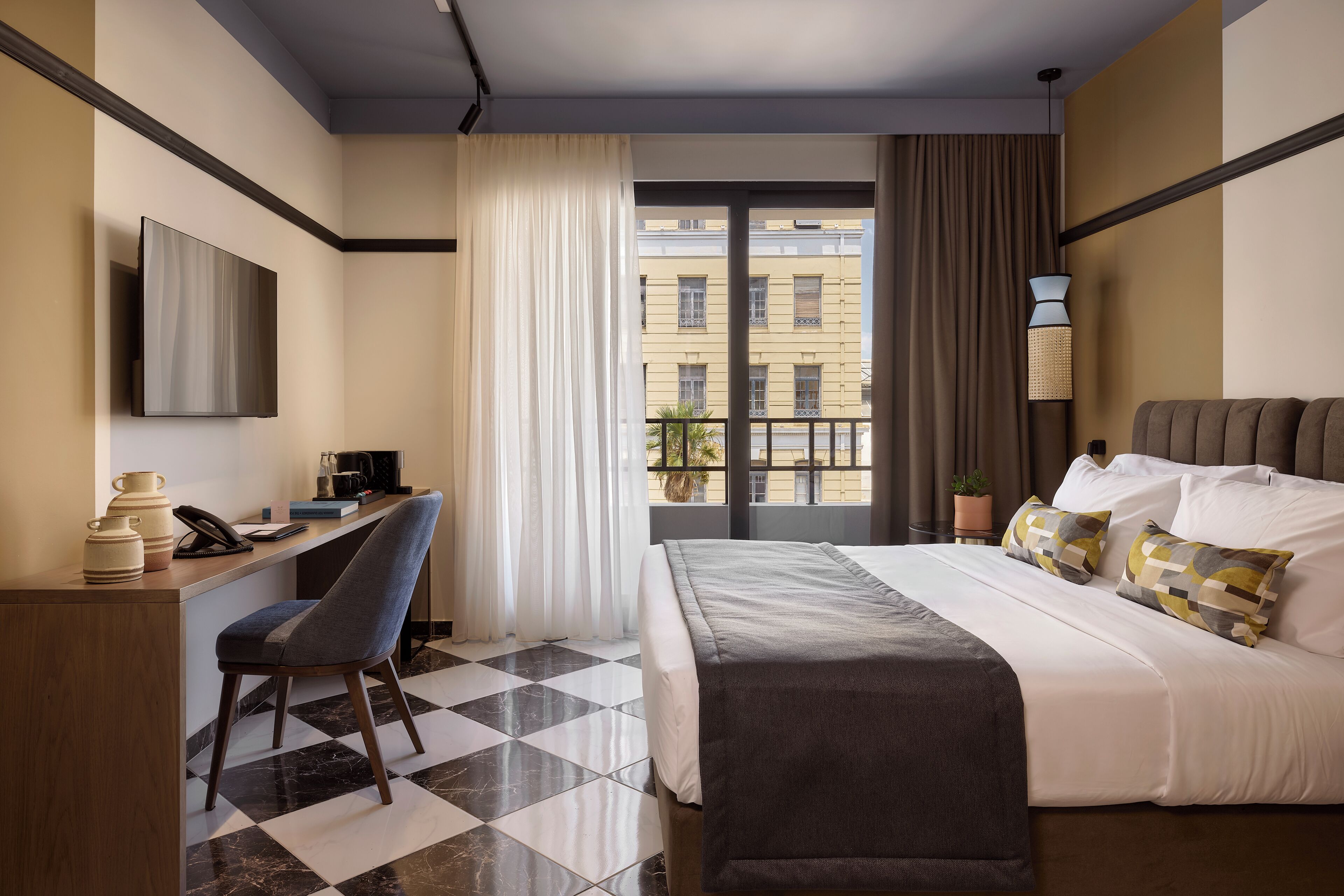 Hotelkamer met een bed, een bureau met stoel, een televisie, een raam met gordijnen en uitzicht op een balkon.