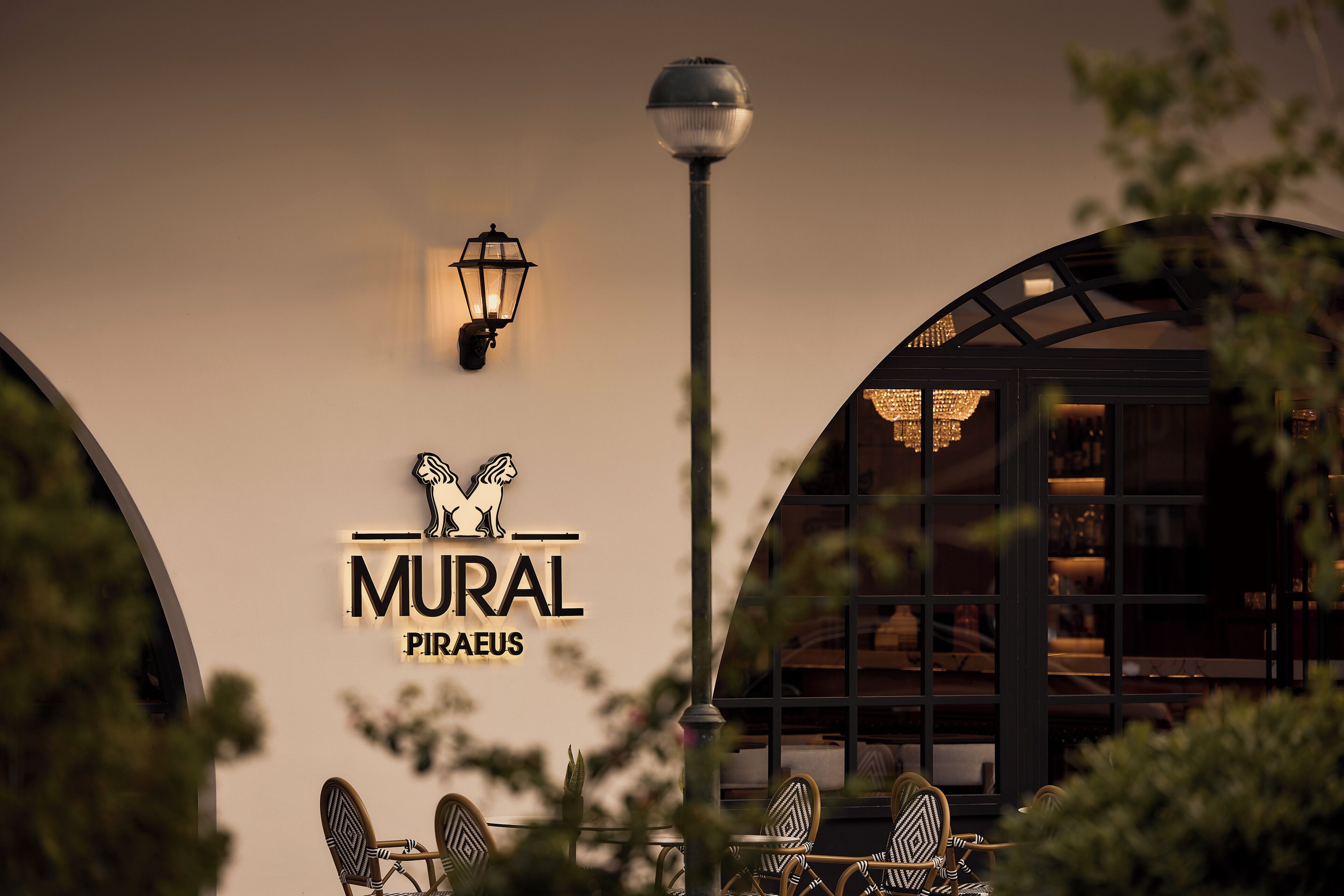 Un restaurant amb un cartell que diu 'MURAL PIRAeus', terrassa amb seients i un fanal.