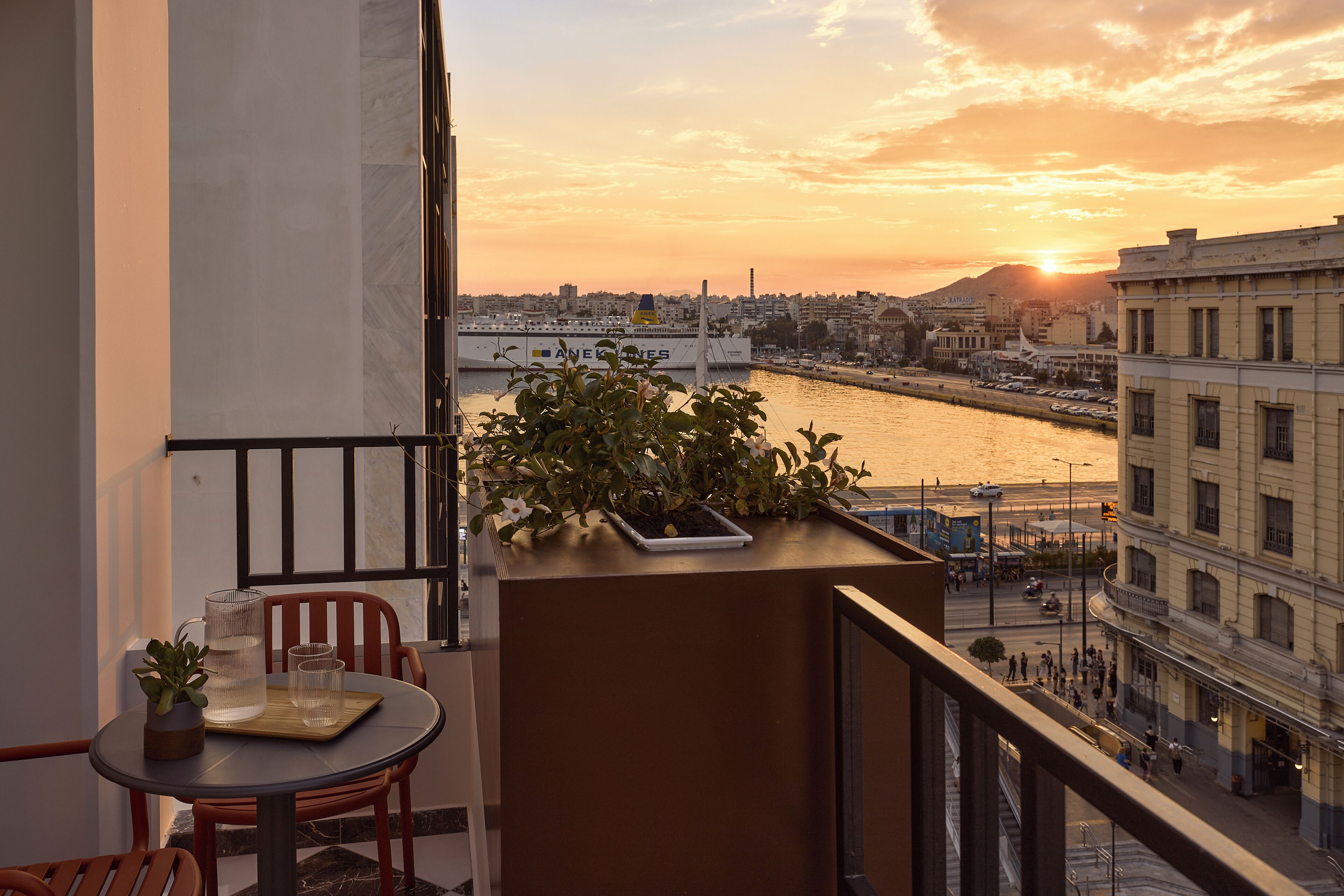 A balcony with a table, chairs, a potted plant, and a view of a river and cityscape at sunset.