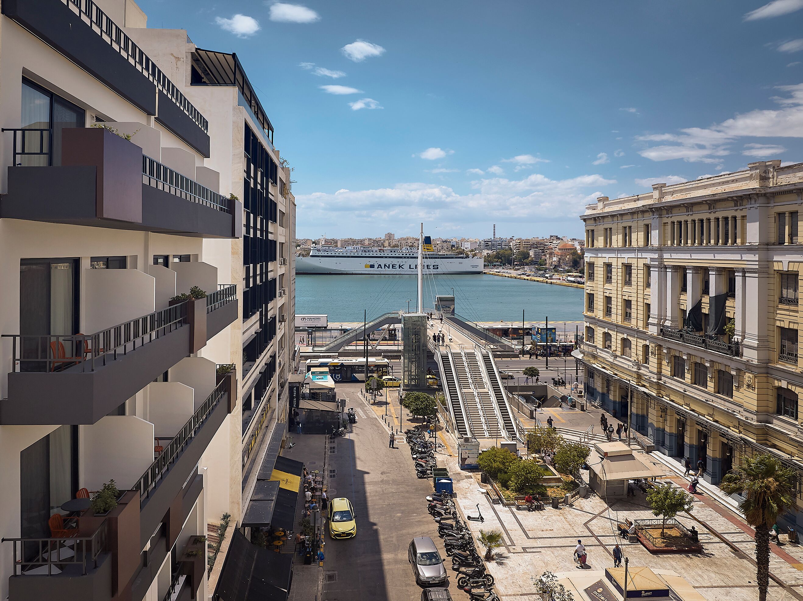 A modern building with balconies, a pedestrian area, and a view of a large cruise ship in the background.