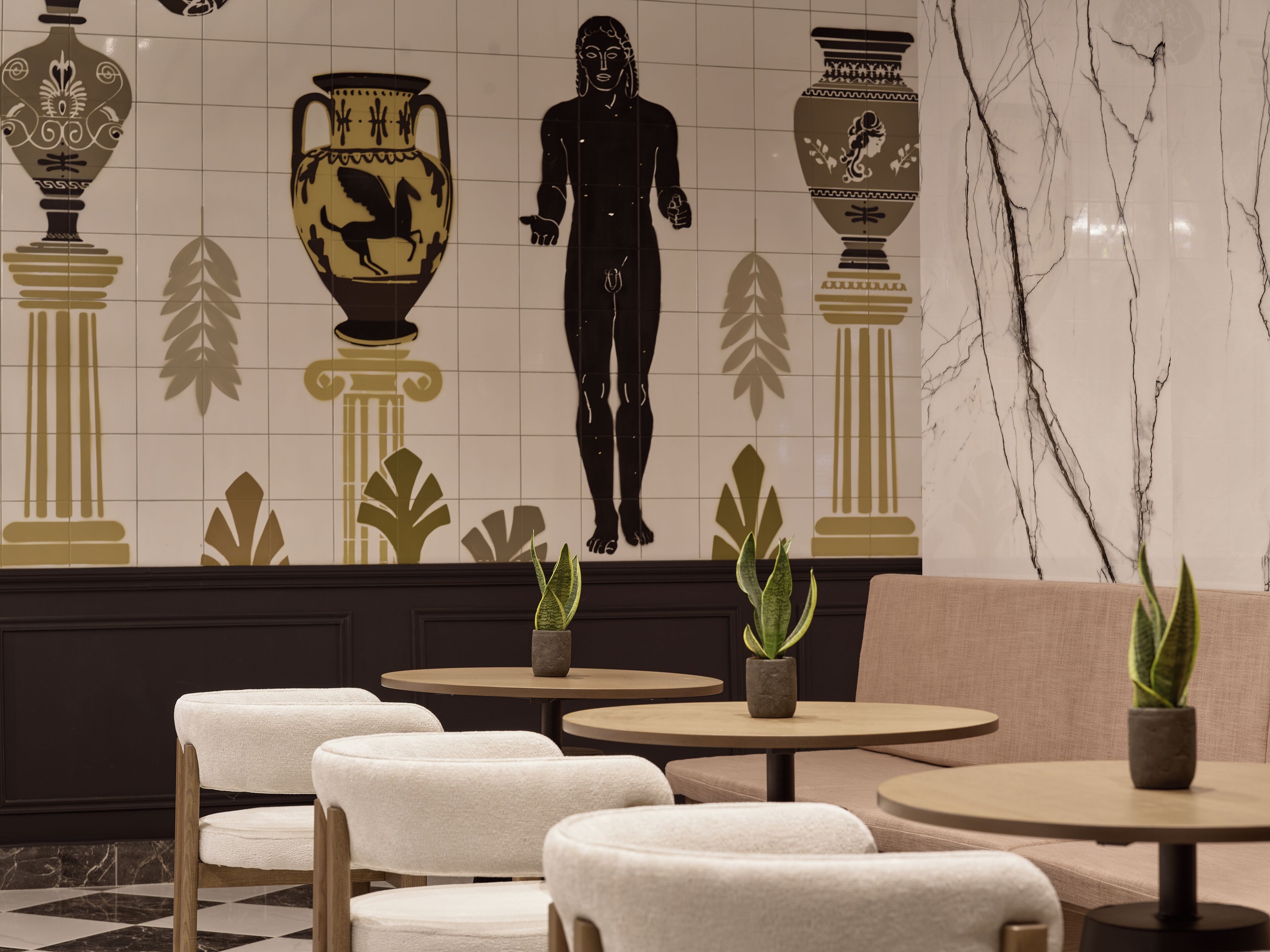 A lobby with a tiled wall featuring classical art, a seating area with round tables and chairs, and potted plants.