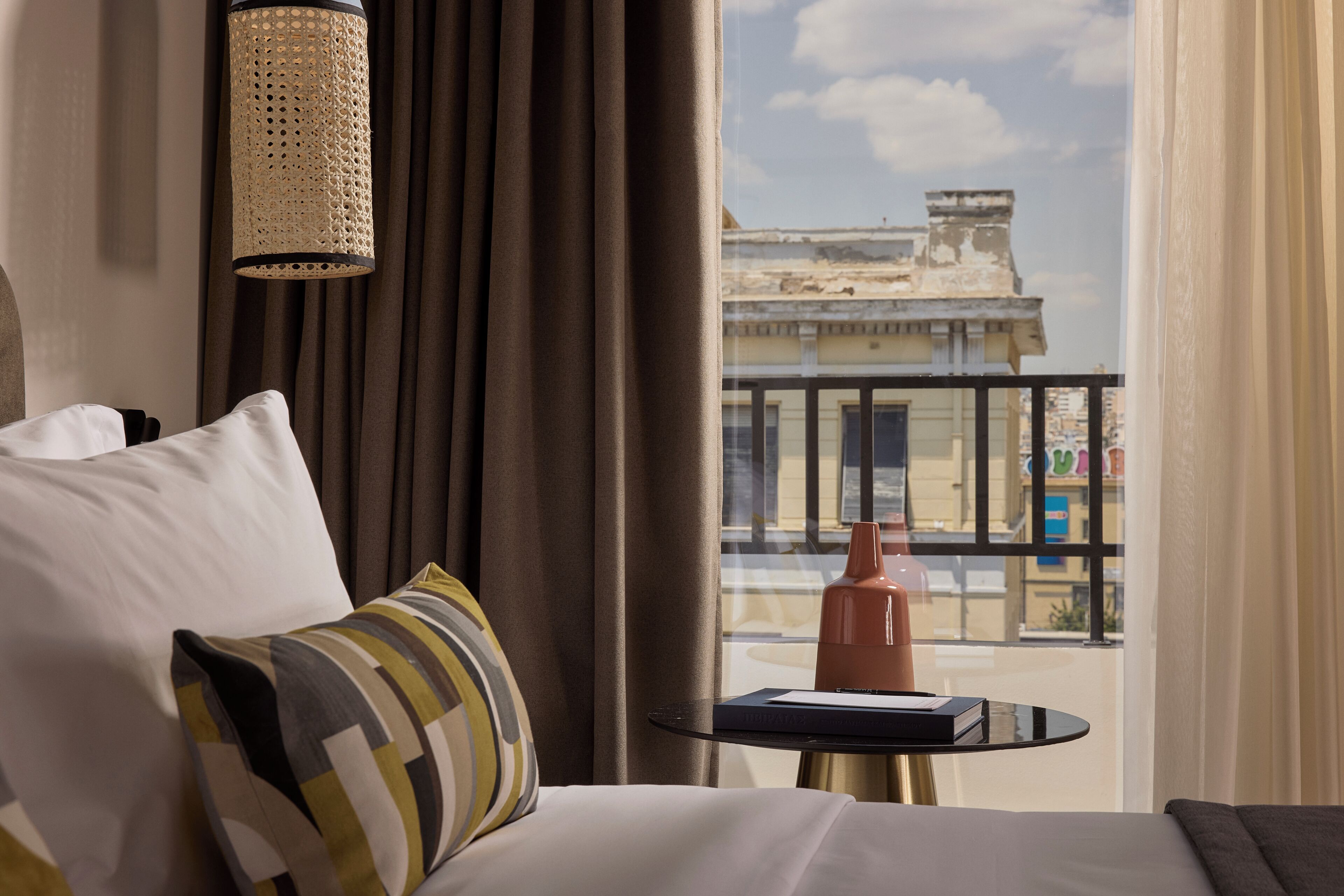 Quarto de hotel com cama, uma mesinha, um abajur e vista para edifícios pela janela.