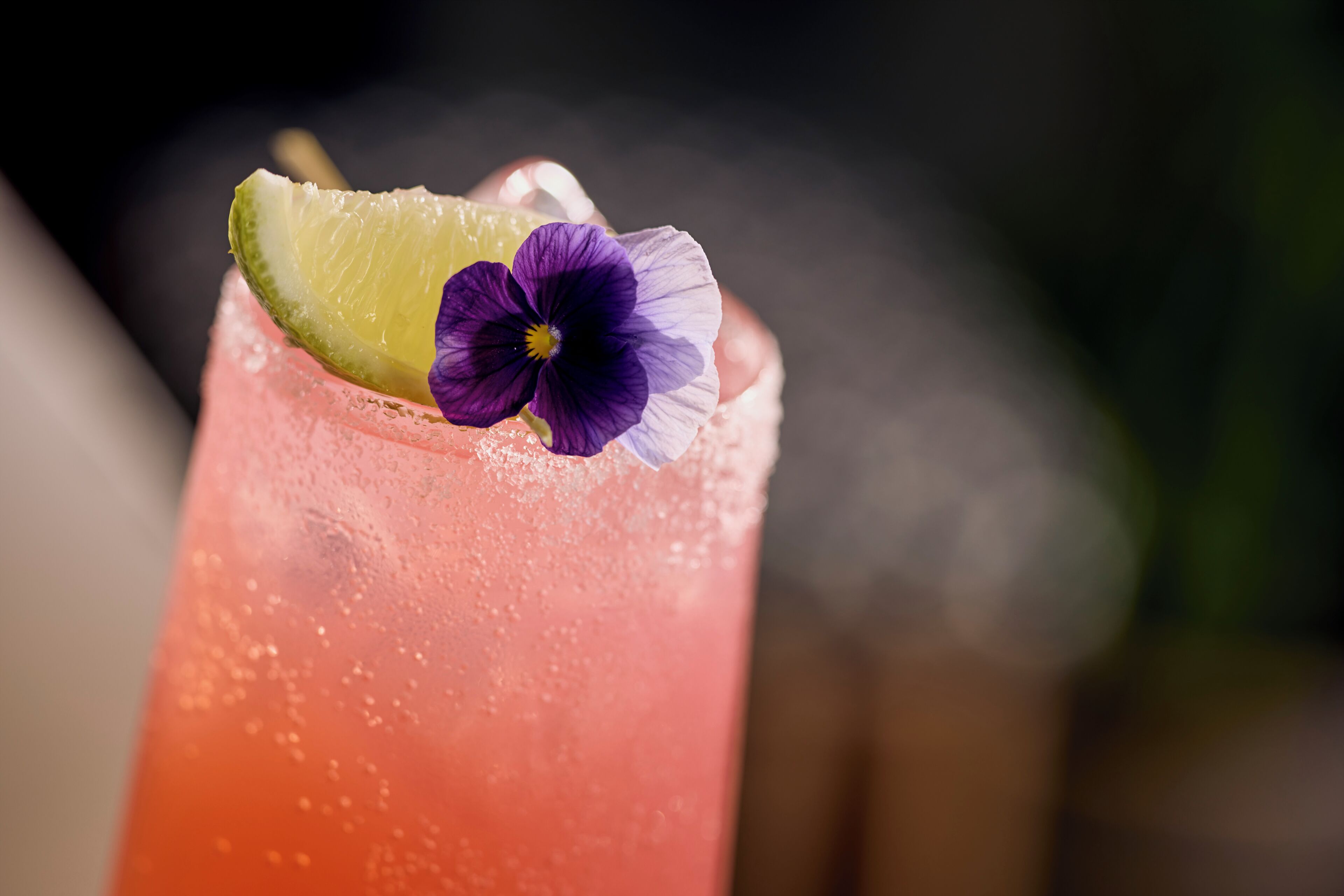 A close-up of a beverage with a lime wedge and a purple flower garnish.