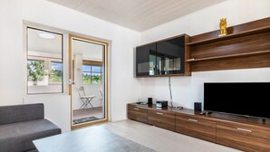A modern living room with a grey sofa, a coffee table, a flat-screen TV mounted on a wooden wall, and a sliding glass door leading to an outdoor area.