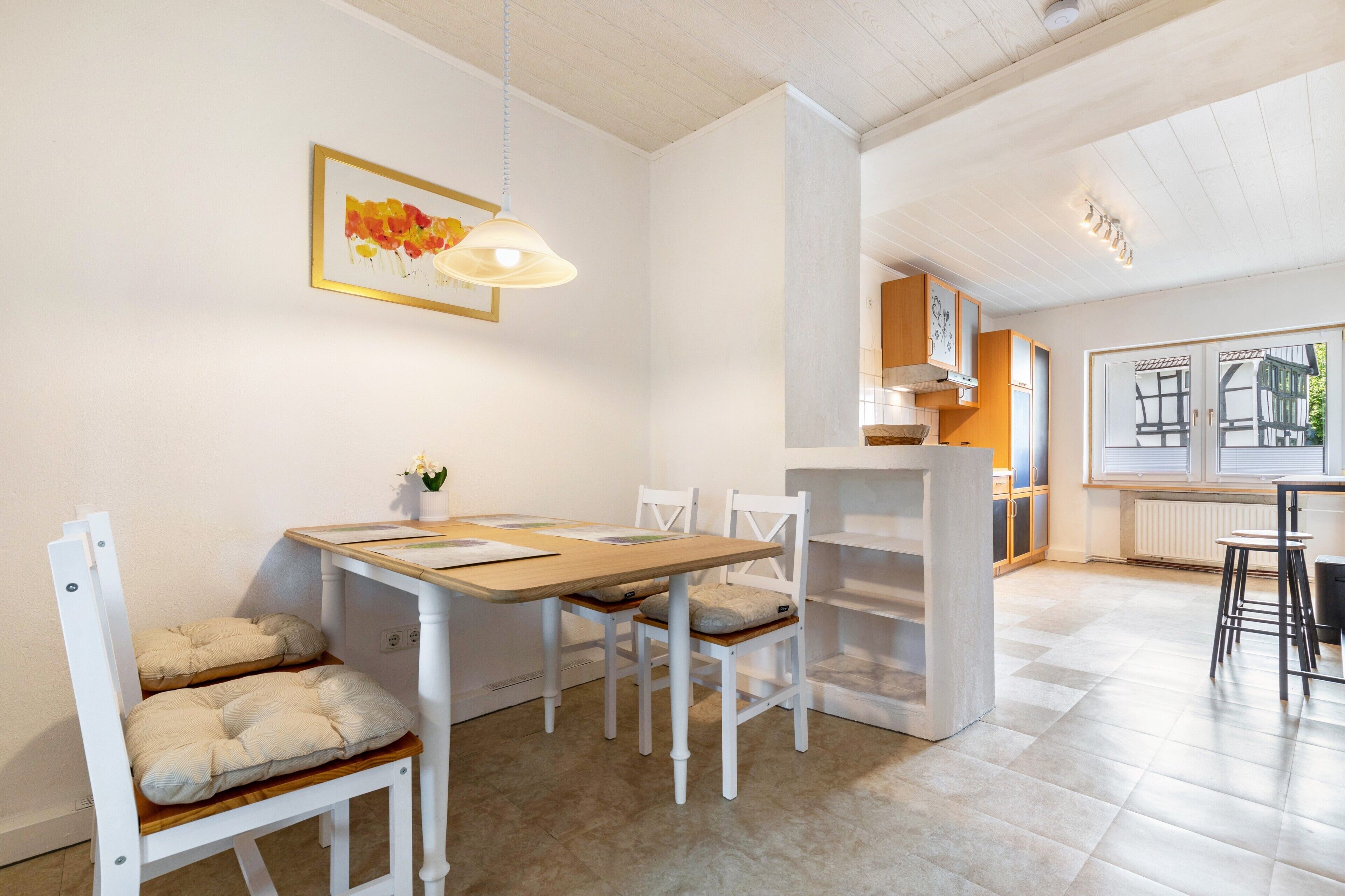 A dining area with a wooden table and white chairs, a pendant light, and a view of a kitchen and a window with blinds.