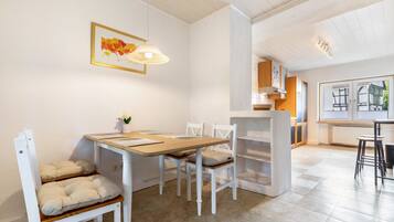A dining area with a wooden table and white chairs, a pendant light, and a view of a kitchen and a window with blinds.