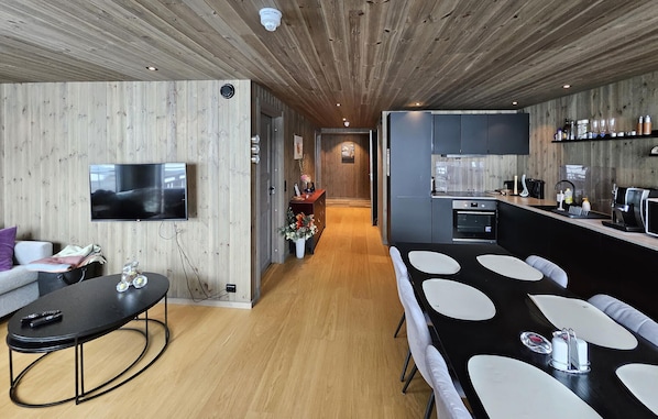 A modern interior with a wooden ceiling, a TV, a dining area with white chairs, a kitchen with dark cabinets, and a hallway leading to another room.