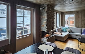 A cozy living room with a stone fireplace, large windows, a sofa, and a dining table.