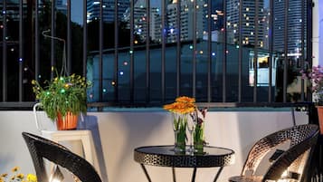 A rooftop terrace with two wicker chairs, a small round table with a vase of flowers, and a view of a city skyline at dusk.