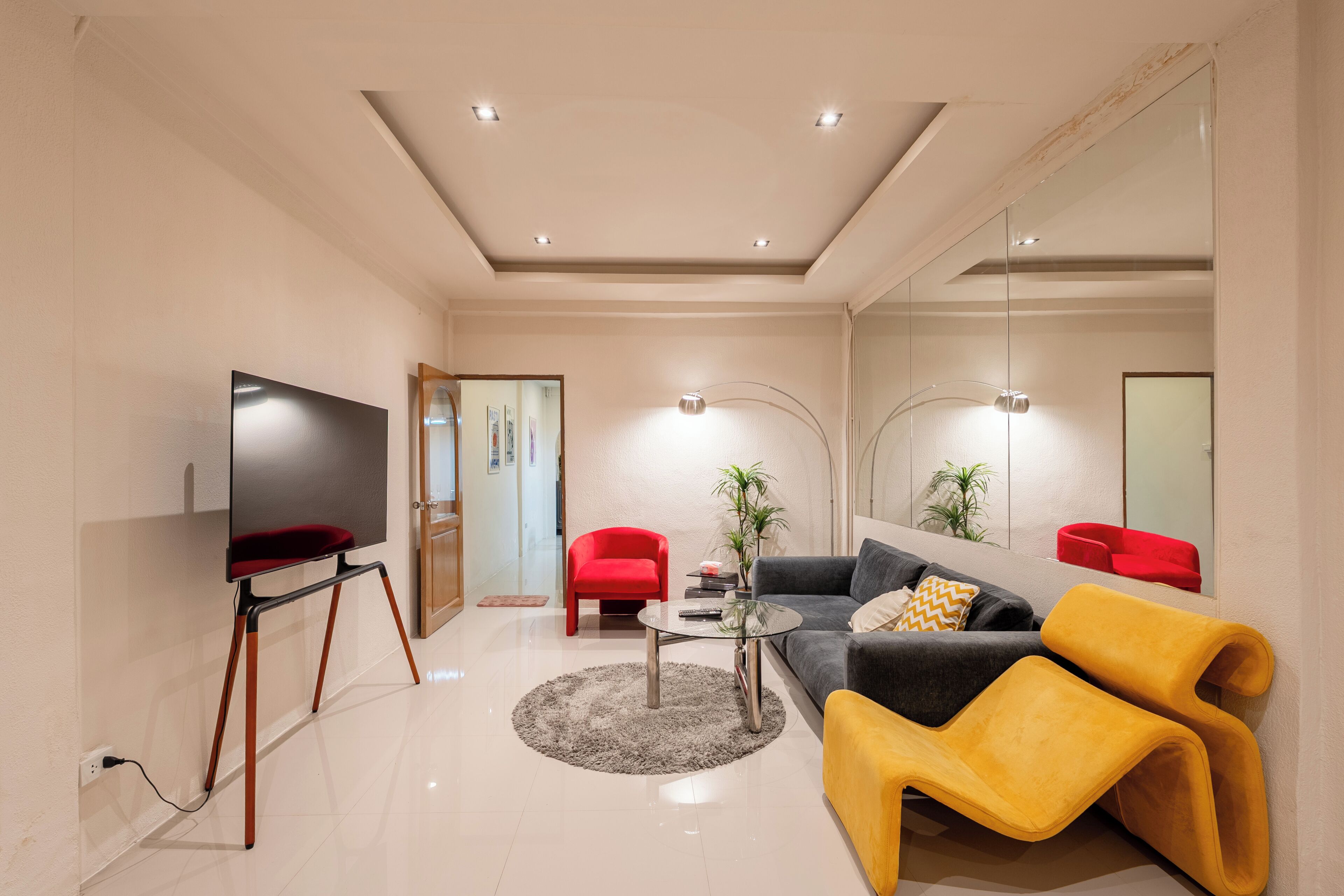 A modern living room with a flat-screen TV, a glass coffee table, a sofa, a yellow armchair, and a red chair.