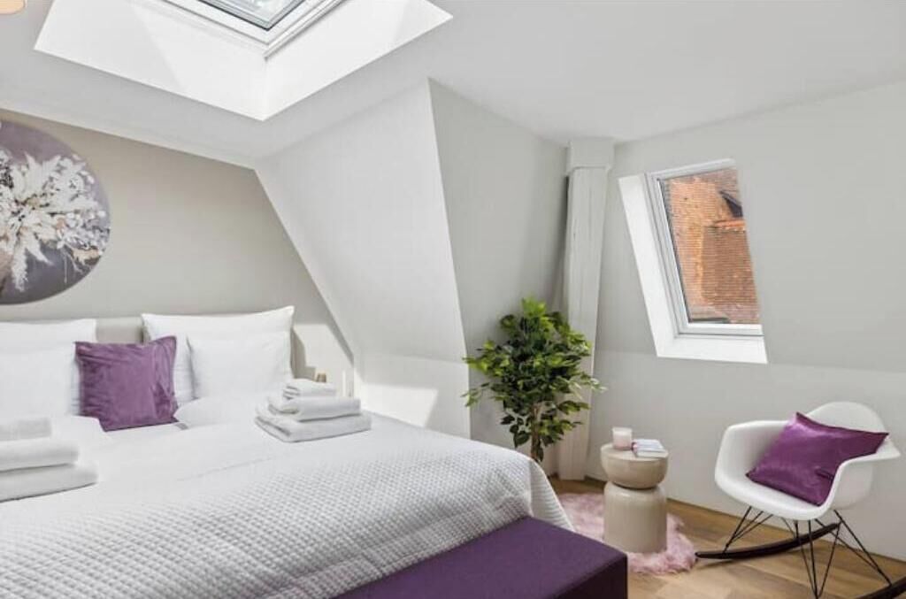 Un dormitorio moderno con tragaluz, una cama con ropa de cama blanca y almohadas moradas, una mesita de noche, una silla y una planta en maceta.